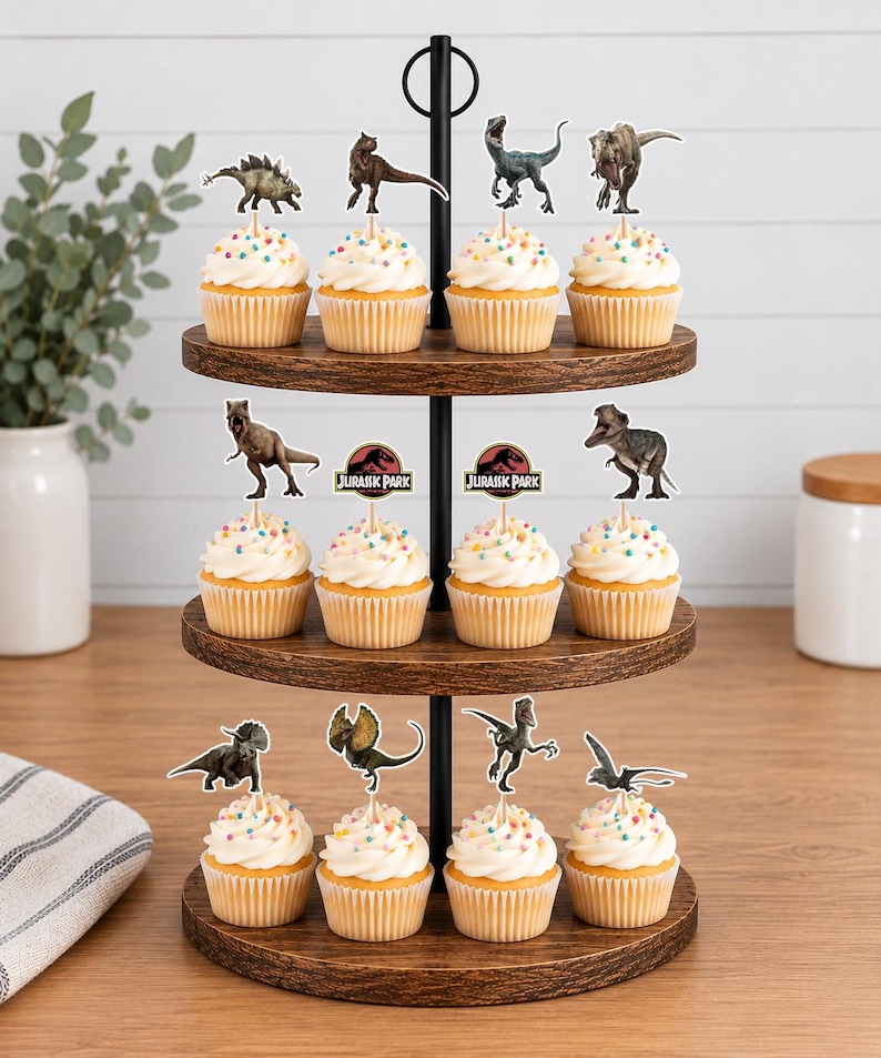 Pu&ograve; includere: Un'alzata in legno a tre ripiani con dodici cupcake guarniti con glassa bianca, zuccherini colorati e vari topper in carta a tema dinosauri, inclusi due loghi circolari di Jurassic Park montati su stecchini di legno.