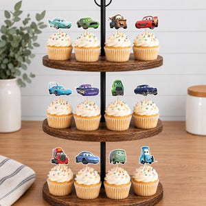 May include: A three-tiered wooden cupcake stand featuring twelve cupcakes topped with colorful sprinkles and individual paper cutouts of various animated car characters from the movie Cars, including Lightning McQueen, Mater, and others.