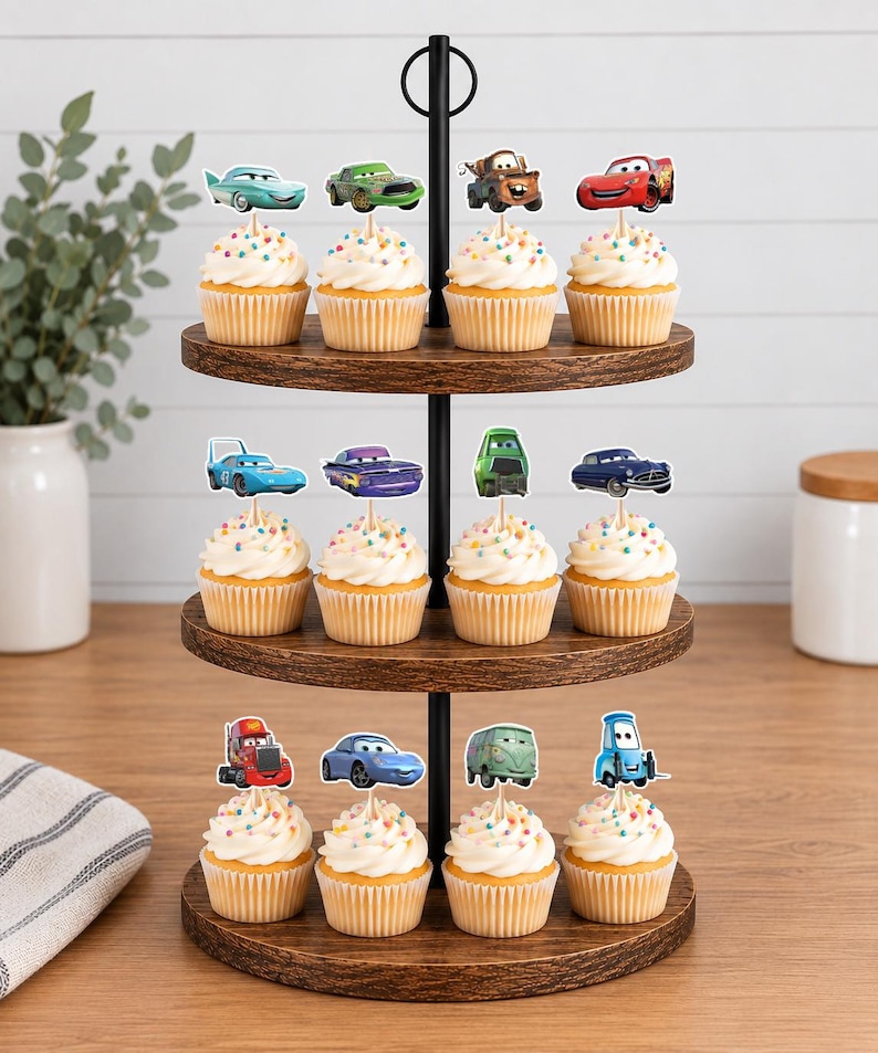 May include: A three-tiered wooden cupcake stand featuring twelve cupcakes topped with colorful sprinkles and individual paper cutouts of various animated car characters from the movie Cars, including Lightning McQueen, Mater, and others.