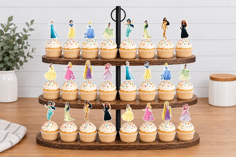 May include: A three-tiered wooden cupcake stand holding twenty-three cupcakes, each topped with a unique paper cutout of a different female character in a colorful gown. The cupcakes feature white frosting with multicolored sprinkles in paper liners.