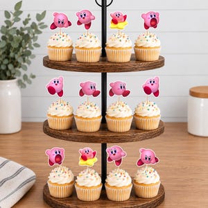 May include: A three-tiered wooden cupcake stand holding twelve cupcakes, each topped with a pink Kirby character paper topper. The cupcakes feature white frosting with colorful sprinkles, and the stand has a black metal central handle.