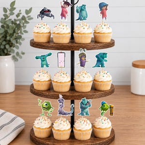 May include: A three-tiered wooden cupcake stand holding twelve cupcakes topped with white frosting and colourful sprinkles. Each cupcake features a paper cutout topper depicting various characters and objects from the movie Monsters, Inc.