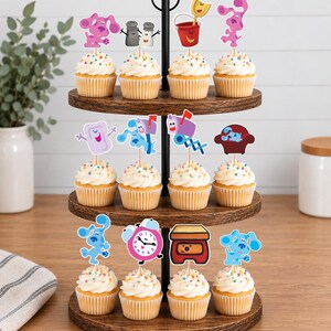 May include: A three-tiered wooden cupcake stand holding twelve cupcakes topped with colorful sprinkles and paper cutouts of Blue's Clues characters and objects, including Magenta, Salt and Pepper, a bucket, a mailbox, a chair, and a clock.