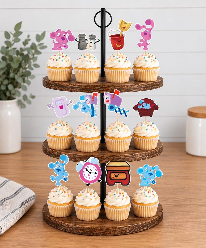 May include: A three-tiered wooden cupcake stand holding twelve cupcakes topped with colorful sprinkles and paper cutouts of Blue's Clues characters and objects, including Magenta, Salt and Pepper, a bucket, a mailbox, a chair, and a clock.