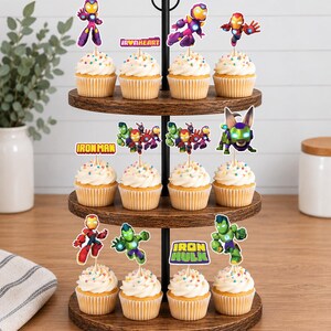 May include: A three-tiered wooden cupcake stand holding twelve cupcakes topped with colorful sprinkles and various superhero-themed paper toppers, including characters like Iron Man, Ironheart, and Iron Hulk, with text labels reading Ironheart, Ironman, and Iron Hulk.