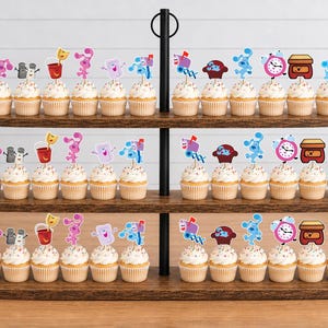 May include: A three-tiered wooden cupcake stand holding numerous cupcakes topped with paper cutouts of Blue's Clues characters and objects, including Blue, Magenta, Mailbox, Shovel and Pail, Tickety Tock, and Side Table Drawer on wooden picks.