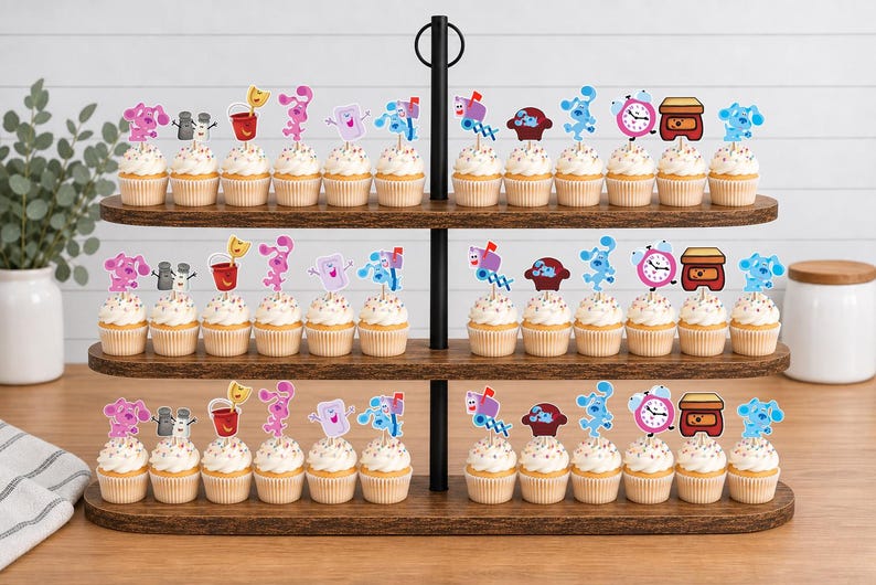 May include: A three-tiered wooden cupcake stand holding numerous cupcakes topped with paper cutouts of Blue's Clues characters and objects, including Blue, Magenta, Mailbox, Shovel and Pail, Tickety Tock, and Side Table Drawer on wooden picks.