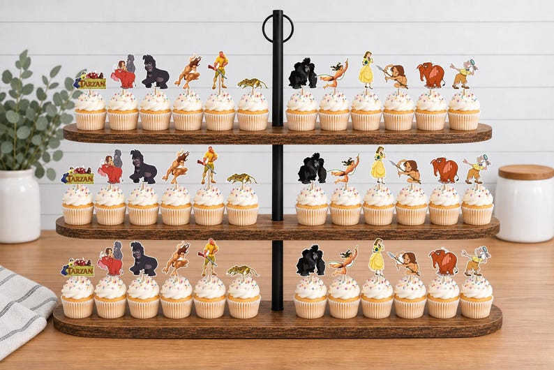 May include: A three-tiered wooden cupcake stand holding numerous cupcakes topped with paper cutouts of Tarzan characters, including Tarzan, Jane, gorillas, and other jungle animals, with a Tarzan logo sign on the left side of each tier.