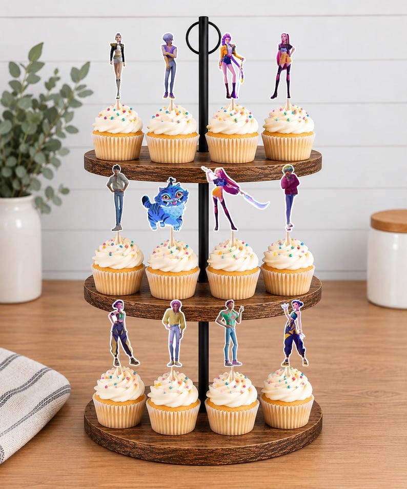 Op de afbeelding: Een houten cupcake-standaard met drie niveaus en twaalf cupcakes, bedekt met witte glazuur en kleurrijke hagelslag. Elke cupcake is voorzien van een decoratieve papieren topper met diverse geanimeerde figuren, waaronder een blauw katachtig wezen.