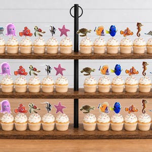 May include: A three-tiered wooden cupcake stand holding rows of cupcakes topped with various sea creature character cutouts, including a shark, octopus, clownfish, sea turtle, angelfish, starfish, blue tang, seahorse, and stingray.