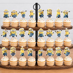 May include: A three-tiered wooden cupcake stand holding twenty-two cupcakes, each topped with a paper Minion character cutout. The cupcakes feature white frosting with colorful sprinkles in light brown paper liners, arranged on dark wood shelves.