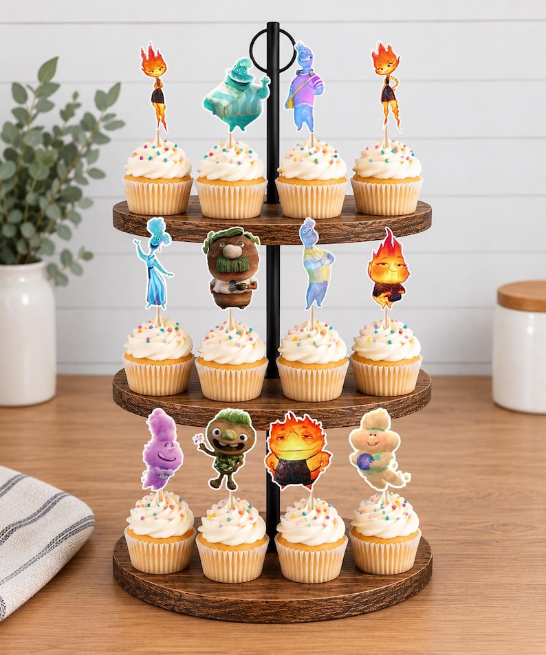 May include: A three-tiered wooden cupcake stand holding twelve cupcakes, each topped with a decorative paper cutout featuring various colourful animated characters from the movie Elemental, including Ember, Wade, and other supporting figures.