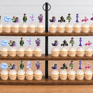 May include: A three-tiered wooden cupcake stand holding rows of cupcakes topped with various character-themed paper toppers. The toppers feature colourful illustrations of diverse figures, creatures, and abstract shapes, each attached to a small pick.