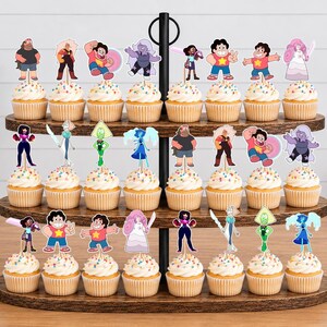 Puede incluir: Un soporte de madera para cupcakes de tres niveles con veinticuatro cupcakes, cada uno decorado con un recorte de papel colorido de un personaje de la serie animada Steven Universe. Los cupcakes tienen glaseado blanco con chispas de colores.