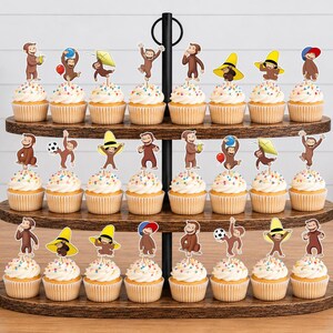 May include: A three-tiered wooden cupcake stand holding twenty-four cupcakes, each topped with a paper cutout of Curious George in various poses, including wearing hats, holding balloons, playing with a soccer ball, or flying a kite.