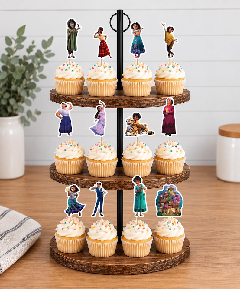 Pode incluir: Um suporte de madeira para cupcakes de tr&ecirc;s n&iacute;veis com doze cupcakes cobertos com cobertura branca e granulado colorido. Cada cupcake possui um topo de papel decorativo retratando v&aacute;rios personagens e uma casa do filme Encanto.
