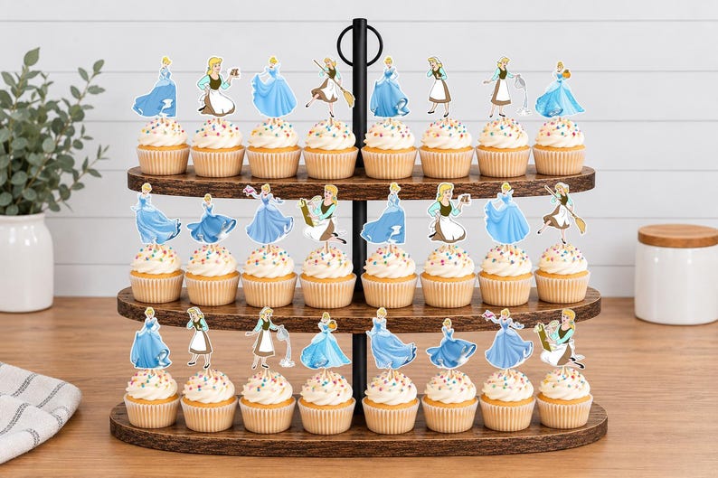 May include: A three-tiered wooden cupcake stand holding twenty-four cupcakes, each topped with a paper cutout of a character in either a blue gown or a brown and white dress. The cupcakes feature white frosting with colorful sprinkles in paper liners.