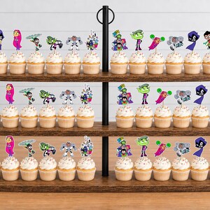 May include: A three-tiered wooden cupcake stand holding numerous cupcakes topped with colourful paper cutouts of Teen Titans characters including Raven, Starfire, Robin, Beast Boy, and Cyborg, each featuring vibrant illustrations on wooden picks.