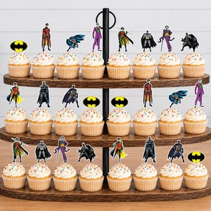 May include: A three-tiered wooden cupcake stand holding twenty-four cupcakes topped with paper cutouts of Batman, Robin, The Joker, and the yellow and black Batman logo. The stand features a central black metal handle and oval-shaped shelves.
