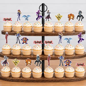K&ouml;nnte beinhalten: Ein dreist&ouml;ckiger Cupcake-St&auml;nder aus Holz mit vierundzwanzig Cupcakes, die mit bunten Streuseln verziert sind. Jeder Cupcake hat einen dekorativen Papieraufsatz mit verschiedenen Yu-Gi-Oh!-Figuren, Symbolen und dem roten Yu-Gi-Oh!-Logo.