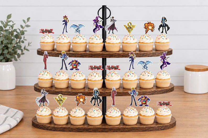 K&ouml;nnte beinhalten: Ein dreist&ouml;ckiger Cupcake-St&auml;nder aus Holz mit vierundzwanzig Cupcakes, die mit bunten Streuseln verziert sind. Jeder Cupcake hat einen dekorativen Papieraufsatz mit verschiedenen Yu-Gi-Oh!-Figuren, Symbolen und dem roten Yu-Gi-Oh!-Logo.