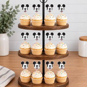 以下が含まれることがあります： 3段の木製カップケーキスタンドに12個のカップケーキが並んでいます。各カップケーキには白いフロスティング、カラフルなスプリンクル、そして中央に挿された木製のピックにミッキーマウスの白黒の顔の紙製カットアウトが飾られています。