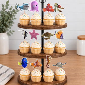 May include: A three-tiered wooden cupcake stand holding twelve cupcakes, each topped with a decorative paper cutout of a sea creature character. The cupcakes feature white frosting with colorful sprinkles and are arranged on circular wooden trays.