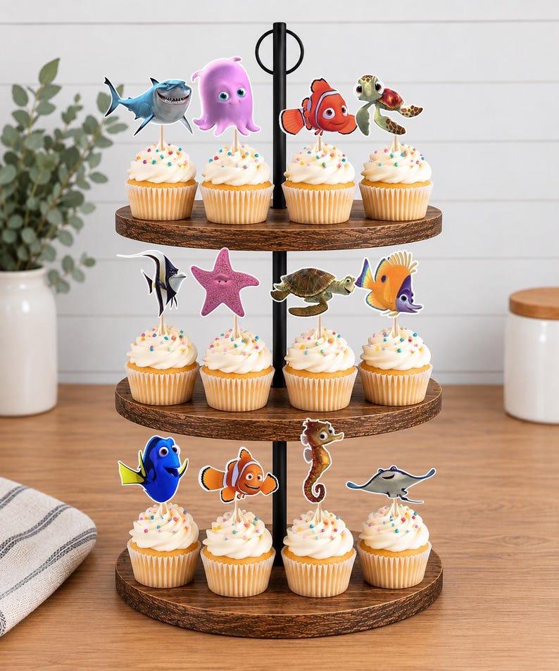 May include: A three-tiered wooden cupcake stand holding twelve cupcakes, each topped with a decorative paper cutout of a sea creature character. The cupcakes feature white frosting with colorful sprinkles and are arranged on circular wooden trays.