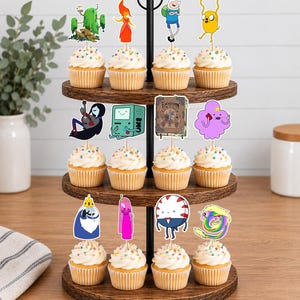 K&ouml;nnte beinhalten: Ein dreist&ouml;ckiger Cupcake-St&auml;nder aus Holz mit zw&ouml;lf Cupcakes, die mit wei&szlig;em Zuckerguss und bunten Streuseln verziert sind. Jeder Cupcake hat einen Papieraufstecker mit verschiedenen Adventure Time-Charakteren wie Finn und Jake.