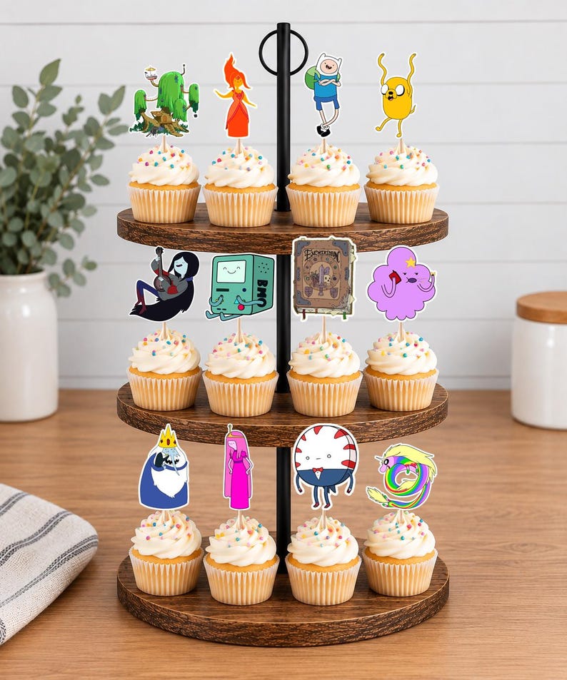 K&ouml;nnte beinhalten: Ein dreist&ouml;ckiger Cupcake-St&auml;nder aus Holz mit zw&ouml;lf Cupcakes, die mit wei&szlig;em Zuckerguss und bunten Streuseln verziert sind. Jeder Cupcake hat einen Papieraufstecker mit verschiedenen Adventure Time-Charakteren wie Finn und Jake.