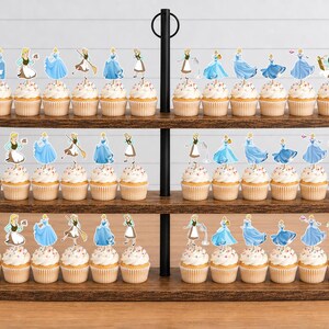 May include: A three-tiered wooden cupcake stand holding numerous cupcakes topped with paper cutouts of a character in various blue dresses and aprons. The cupcakes are arranged in rows on the dark wood shelves, featuring white frosting and light-colored liners.