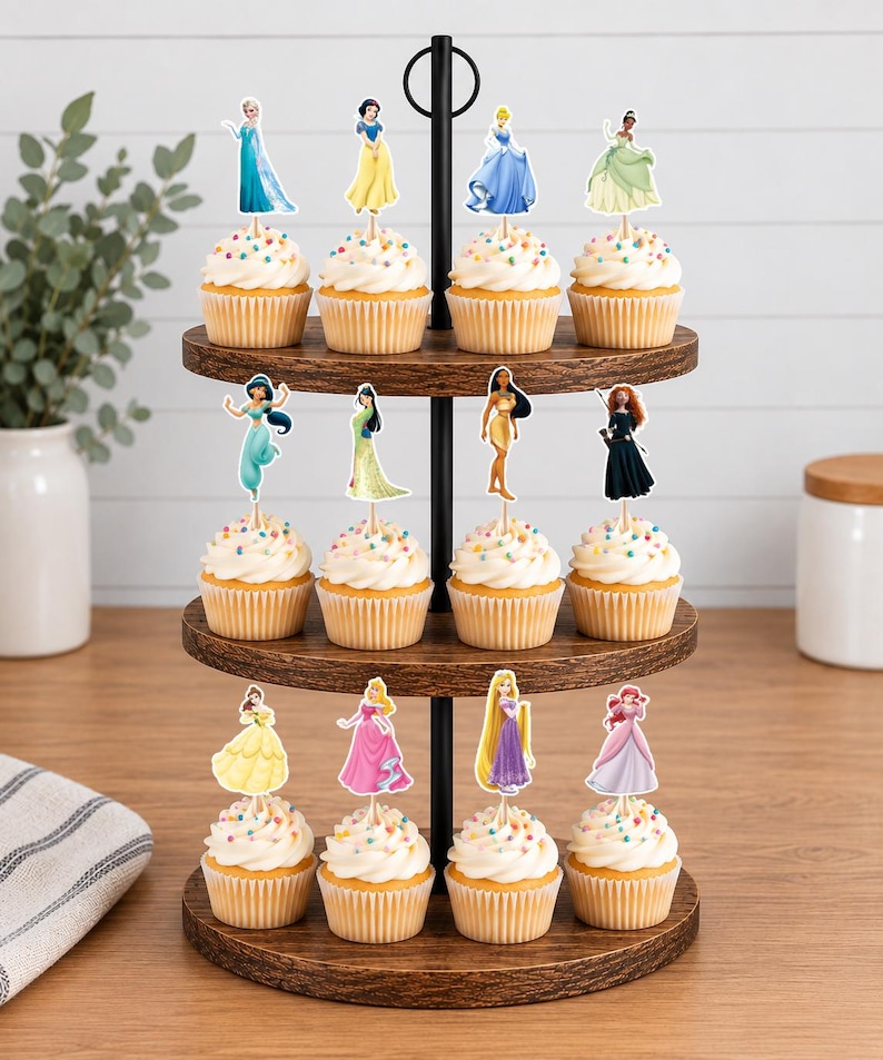 May include: A three-tiered wooden cupcake stand holding twelve cupcakes, each topped with a decorative paper cutout of a different animated princess character. The stand features a central black metal rod and circular wooden shelves with a dark wood grain finish.