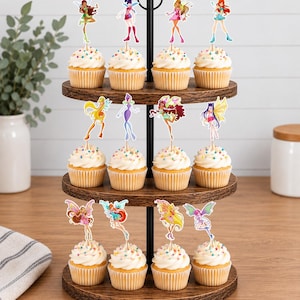 May include: A three-tiered wooden cupcake stand holding twelve cupcakes, each topped with a white frosting swirl, colorful sprinkles, and a decorative paper cutout of a Winx Club fairy character attached to a thin wooden pick.