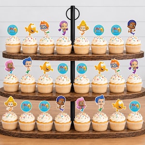 May include: A three-tiered dark wood cupcake stand holding twenty-four cupcakes topped with colorful paper toppers featuring cartoon characters and circular blue logos with the text Bubble Guppies in a variety of poses and expressions.