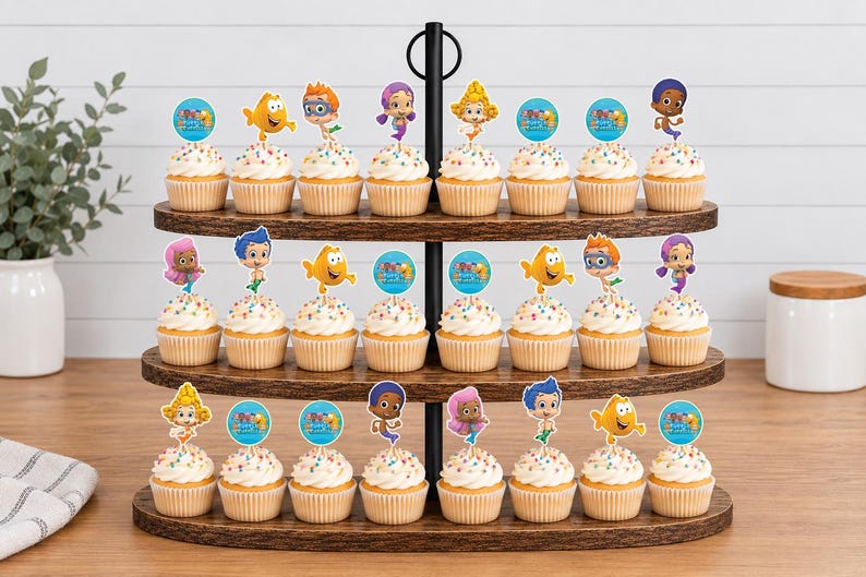 May include: A three-tiered dark wood cupcake stand holding twenty-four cupcakes topped with colorful paper toppers featuring cartoon characters and circular blue logos with the text Bubble Guppies in a variety of poses and expressions.
