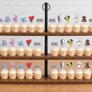 Peut inclure: Un pr&eacute;sentoir &agrave; cupcakes en bois &agrave; trois niveaux contenant des cupcakes surmont&eacute;s de d&eacute;corations en papier repr&eacute;sentant les personnages BT21. Chaque d&eacute;coration affiche un personnage color&eacute; et le logo BT21. Le support a un m&acirc;t central noir.