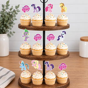 Op de afbeelding: Een houten cupcake-standaard met drie niveaus met twaalf cupcakes, gegarneerd met witte glazuur, kleurrijke hagelslag en papieren My Little Pony-toppers. De toppers tonen verschillende pony's en het My Little Pony-logo op houten prikkers.