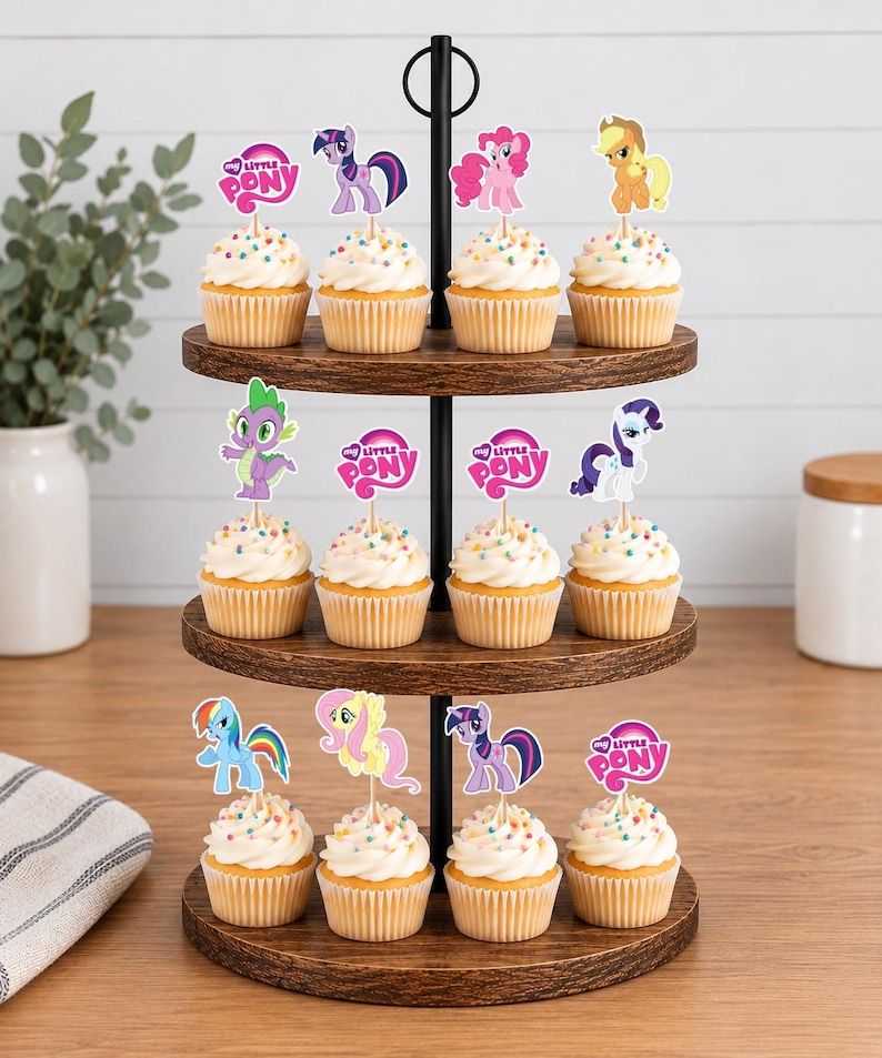 Op de afbeelding: Een houten cupcake-standaard met drie niveaus met twaalf cupcakes, gegarneerd met witte glazuur, kleurrijke hagelslag en papieren My Little Pony-toppers. De toppers tonen verschillende pony's en het My Little Pony-logo op houten prikkers.