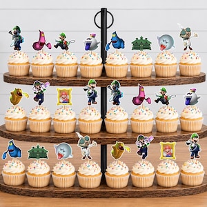 May include: A three-tiered wooden cupcake stand holding twenty-four cupcakes, each topped with a colorful paper cutout featuring characters and icons from the Luigi's Mansion video game series, including Luigi, ghosts, and framed portraits.