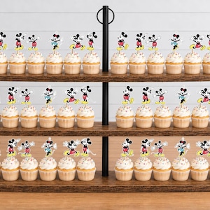 K&ouml;nnte beinhalten: Ein dreist&ouml;ckiger Cupcake-St&auml;nder aus Holz mit zahlreichen Cupcakes, die mit wei&szlig;em Zuckerguss und Papieraufsteckern verziert sind, welche verschiedene Illustrationen von Mickey und Minnie Mouse in den klassischen Farben Rot, Schwarz, Gelb und Wei&szlig; zeigen.