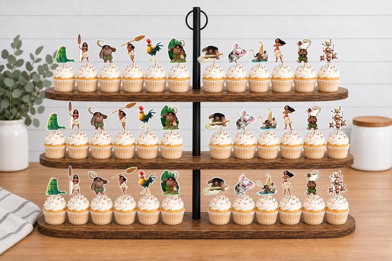Op de afbeelding: Een houten cupcake-standaard met drie niveaus vol cupcakes met papieren decoraties in Moana-thema. De prikkers tonen diverse personages waaronder Moana, Maui, het varkentje Pua, de haan Heihei en andere iconen uit de bekende animatiefilm.
