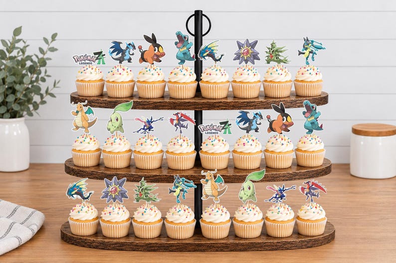 K&ouml;nnte beinhalten: Ein dreist&ouml;ckiger Cupcake-St&auml;nder aus dunklem Holz mit vierundzwanzig Cupcakes, die mit bunten Streuseln und verschiedenen Pok&eacute;mon-Papieraufsteckern, einschlie&szlig;lich des Pok&eacute;mon Legends-Logos, auf drei ovalen Holzablagen dekoriert sind.
