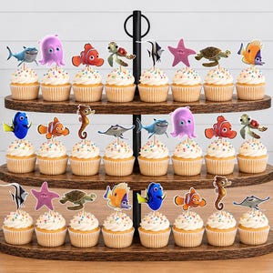 May include: A three-tiered wooden cupcake stand holding twenty-one cupcakes, each topped with a colorful paper cutout of various sea creatures including a blue tang, clownfish, sea turtle, octopus, seahorse, stingray, and starfish characters.