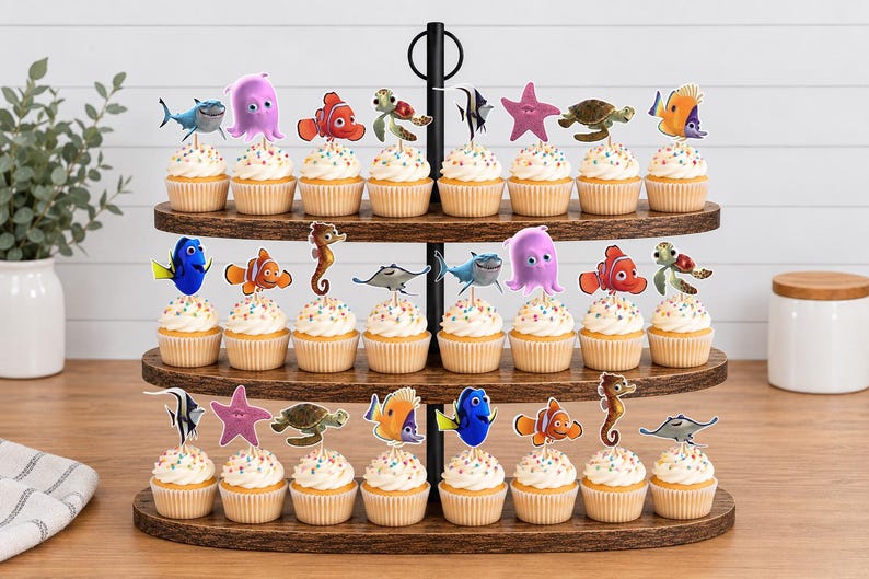 May include: A three-tiered wooden cupcake stand holding twenty-one cupcakes, each topped with a colorful paper cutout of various sea creatures including a blue tang, clownfish, sea turtle, octopus, seahorse, stingray, and starfish characters.
