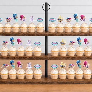May include: A three-tiered wooden cupcake stand holding numerous cupcakes topped with Shimmer and Shine character paper toppers. Each topper features colorful cartoon figures or the Shimmer and Shine logo. The cupcakes have white frosting.