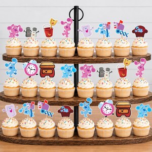 May include: A three-tiered wooden cupcake stand holding twenty-four cupcakes, each topped with a colorful paper cutout featuring characters and objects from Blue's Clues, including Blue, Magenta, a mailbox, a clock, a soap bar, and a red bucket.