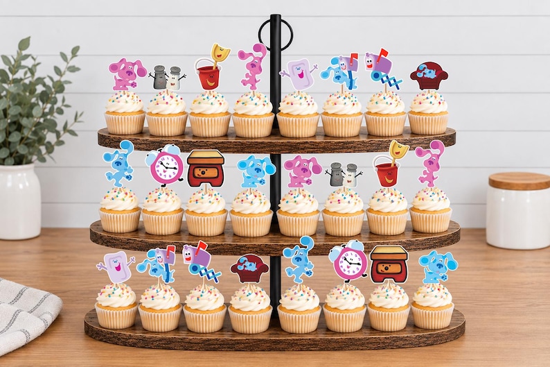 May include: A three-tiered wooden cupcake stand holding twenty-four cupcakes, each topped with a colorful paper cutout featuring characters and objects from Blue's Clues, including Blue, Magenta, a mailbox, a clock, a soap bar, and a red bucket.