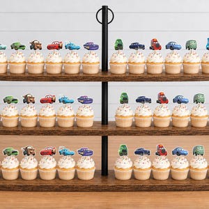 May include: A three-tiered wooden cupcake stand holding numerous cupcakes topped with colorful paper cutouts of various cartoon cars. Each cupcake features white frosting and a unique character topper, arranged in rows on the dark brown wooden shelves.