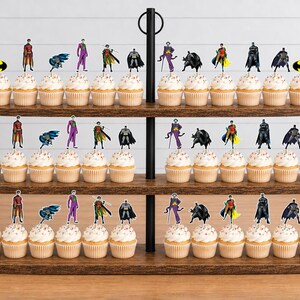 May include: A three-tiered wooden cupcake stand holding numerous cupcakes topped with decorative paper cutouts featuring Batman, Robin, Joker, and the yellow Batman logo. The stand has a black metal central handle and a dark wood finish.