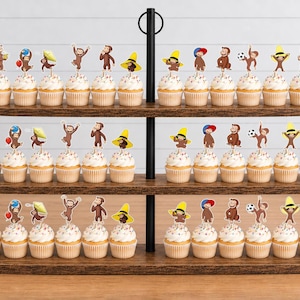 May include: A three-tiered wooden cupcake stand holding numerous cupcakes topped with Curious George character paper toppers. The brown monkey figures are depicted in various poses, some wearing yellow hats or holding items like balloons and soccer balls.